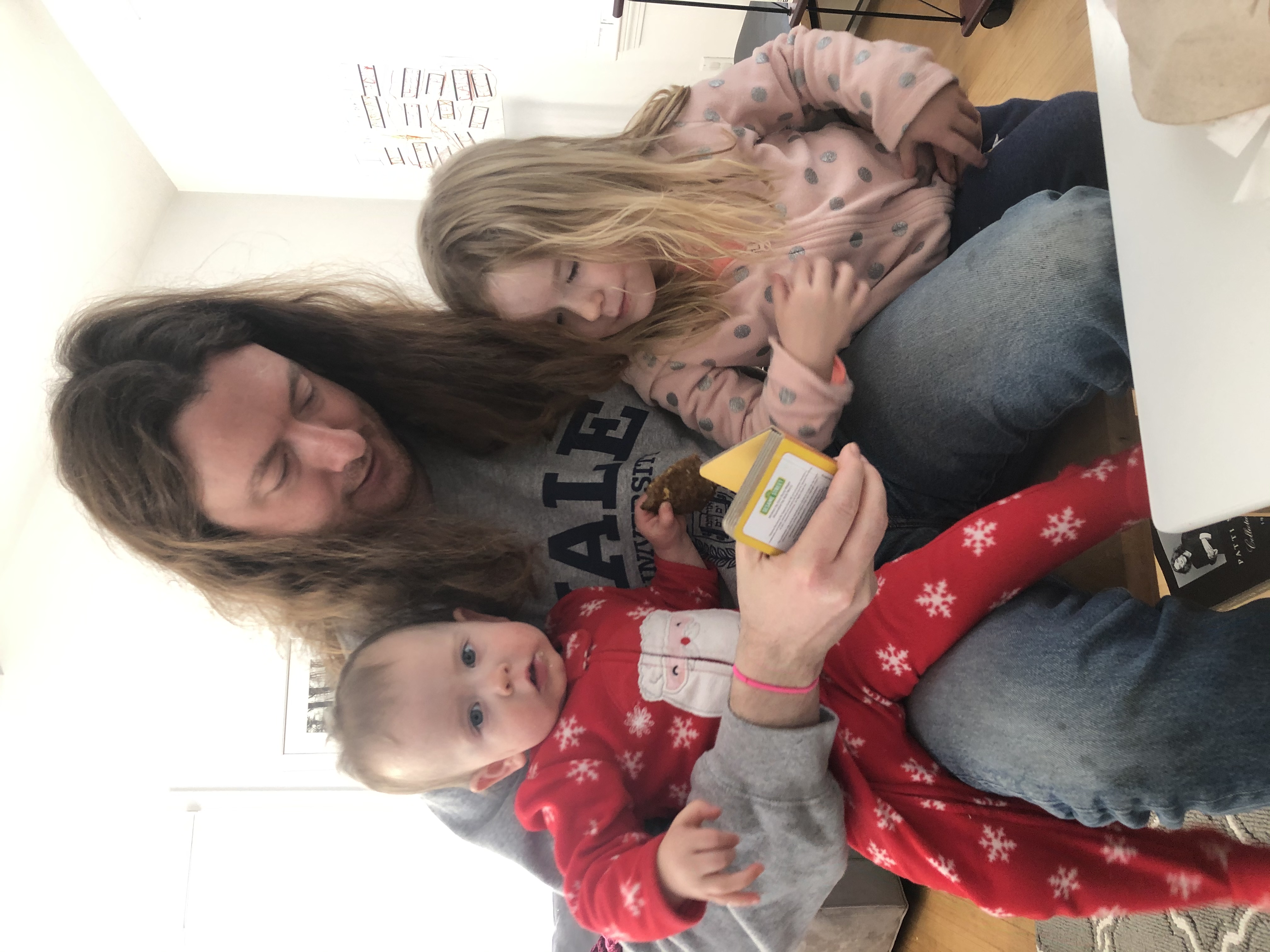 A longhaired dude reading to a 1-year-old and three-year-old.